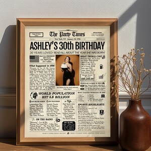 May include: A framed newspaper-style poster titled The Party Times, featuring the headline ASHLEY'S 30TH BIRTHDAY. The print includes a central photograph of a person holding a cake, surrounded by 1996 trivia, statistics, and pop culture facts.