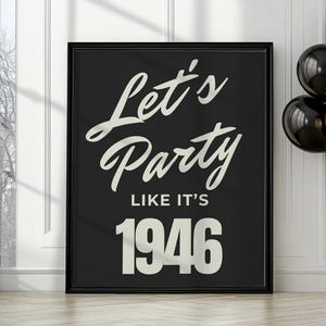 May include: A framed black poster with the text "Let's Party Like It's 1946" in a white, cursive font. The poster is propped against a white wall, with black and gold balloons to the right.