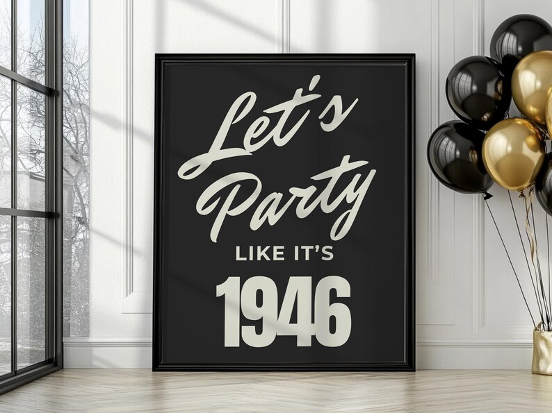 May include: A framed black poster with the text "Let's Party Like It's 1946" in a white, cursive font. The poster is propped against a white wall, with black and gold balloons to the right.
