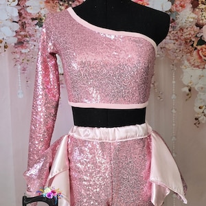 May include: A pink sequined dance costume set, including a one-shoulder crop top and shorts with satin accents. The costume is displayed on a black mannequin. The logo "Thee Quilted Charm" is visible at the bottom.