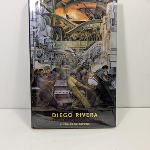 May include: A hardcover book titled Diego Rivera: The Detroit Industry Murals by Linda Bank Downs. The cover features a detailed, colorful mural illustration depicting industrial factory workers operating heavy machinery in a large manufacturing plant.