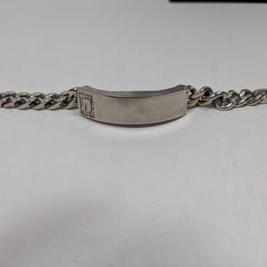May include: A silver-toned bracelet with a curb chain design. The bracelet features a rectangular center plate with a small, square design embellished with small, clear stones. The bracelet is displayed on a white surface.