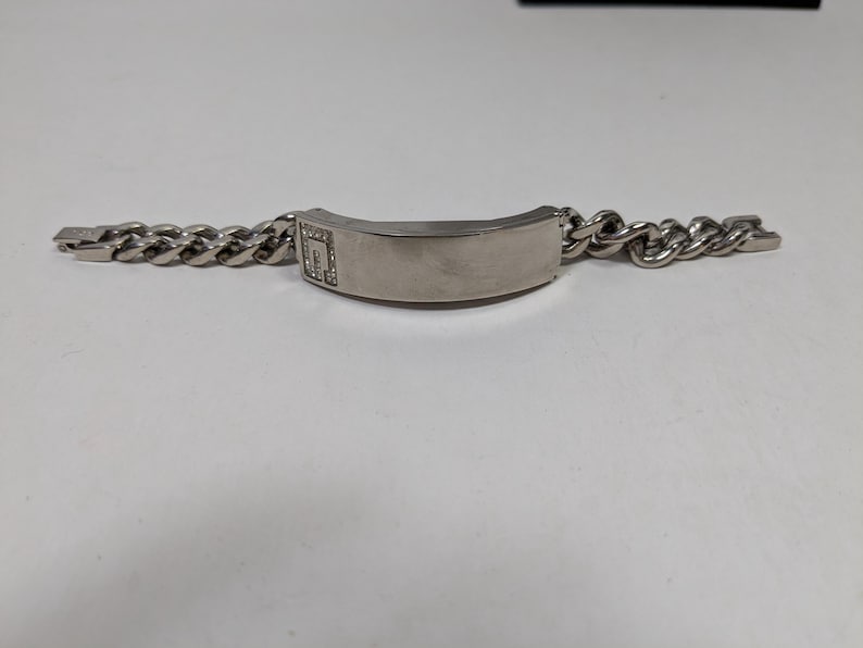 May include: A silver-toned bracelet with a curb chain design. The bracelet features a rectangular center plate with a small, square design embellished with small, clear stones. The bracelet is displayed on a white surface.
