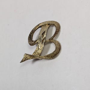 May include: A vintage, metallic letter "B" in a script font. The letter is a tarnished gold color with a textured surface, set against a plain white background. The design is a single, stylized initial.