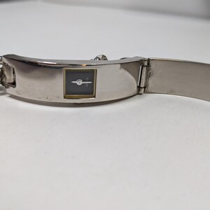May include: A silver-toned wristwatch with a rectangular black watch face and gold-colored trim. The watch has a chain-link band on one side and a hinged, flat band on the other. The watch is displayed on a white surface.