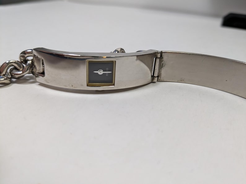 May include: A silver-toned wristwatch with a rectangular black watch face and gold-colored trim. The watch has a chain-link band on one side and a hinged, flat band on the other. The watch is displayed on a white surface.