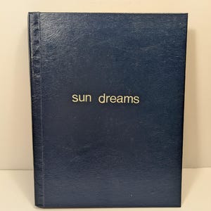 May include: A dark navy blue hardcover book with a textured, leather-like finish. The words "sun dreams" are printed in a simple, gold-colored sans-serif font centered on the front cover. The book features a vertical spine detail on the left side.