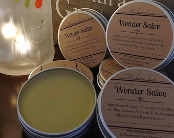 May include: Close-up of three small, round, metal tins containing Wonder Salve. Each tin has a brown label with white text listing the ingredients: Organic Coconut oil & Olive oil, Raw beeswax, Vitamin E oil, Essential oils: Lavender, Tea tree, Lemon. The label also includes the text "Find Mykaproducts @ Facebook, Etsy, Pinterest, Corona 951-741-6102".