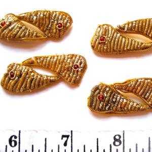 May include: Close-up of several gold-coloured, embroidered appliques. Each applique features a unique shape with a central red jewel. The appliques are arranged on a white surface, with a ruler visible at the bottom.