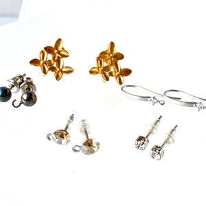 May include: An assortment of earrings in various styles. Includes gold-colored floral stud earrings, silver-toned hook earrings with a single clear stone, and stud earrings with clear and iridescent stones. The earrings are displayed on a white surface.