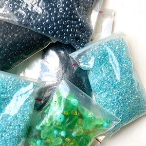 Puede incluir: Cinco bolsas de cuentas de varios colores, incluyendo azul, verde y turquesa. Las cuentas están empaquetadas en bolsas de plástico transparentes con cierres de cremallera rojos.
