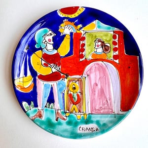 May include: A hand-painted ceramic plate with a vibrant, whimsical design. The plate features a blue rim and a central scene with figures, a red structure, and a fish. The artwork is signed "CANUSA".