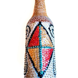 Puede incluir: Una botella de vidrio cubierta de lentejuelas de colores, formando un patrón geométrico. La botella tiene una tapa roja y está decorada con una variedad de colores, incluyendo oro, plata, rojo, verde y azul.