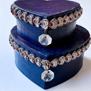 May include: Two-tiered heart-shaped box in deep blue. The box is adorned with a silver-toned trim featuring heart-shaped accents with red and blue jewels. Each tier has a clear, faceted crystal pendant.