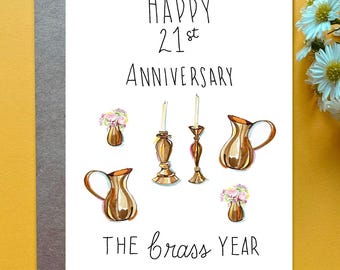 May include: A white greeting card with the text Happy 21st Anniversary The Brass Year. The card features watercolor illustrations of two brass pitchers, two brass candlesticks with white candles, and two small vases with pink flowers.