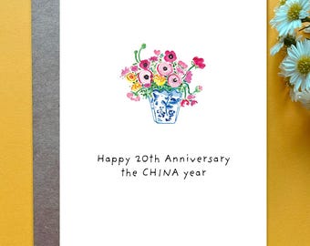 May include: A white greeting card featuring a watercolor illustration of a bouquet of pink, red, and yellow flowers in a blue and white patterned vase. Below the illustration, the text reads: Happy 20th Anniversary the CHINA year.