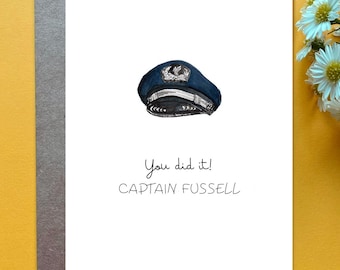 May include: A white greeting card featuring a watercolor illustration of a dark blue pilot cap with a silver emblem. Below the cap, the text reads "You did it!" in a cursive font and "CAPTAIN FUSSELL" in a simple, clean, uppercase font.