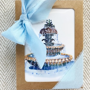 Puede incluir: Una pintura a la acuarela de una fuente escalonada, presentada en una tarjeta marrón y atada con una cinta azul claro. La cinta tiene bordes deshilachados. La fuente presenta agua en cascada. La presentación general es elegante.