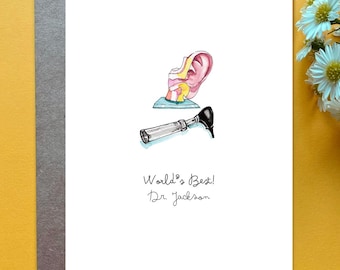 May include: A white greeting card featuring a watercolor illustration of a pink human ear anatomy model and a black otoscope. Below the illustrations, the text reads World's Best! Dr. Jackson in black ink. The card is placed on a gray envelope.