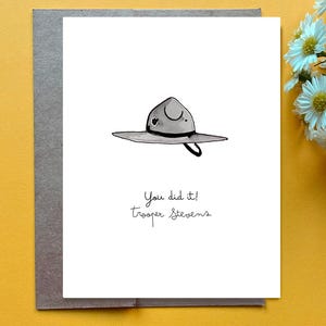 May include: Greeting card with a hand-drawn illustration of a ranger hat. The text "You did it! Trooper Stevens" is written below the hat in a handwritten font. The card is on a gray envelope, set against a yellow background.