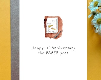 May include: A white greeting card featuring a watercolor illustration of a document with two gold keys and a pen, with the text: Happy 1st Anniversary the PAPER year. The card is placed on a gray envelope against a yellow background.