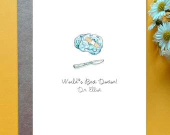 May include: A white greeting card featuring a watercolor illustration of a light blue brain with a tan adhesive bandage and a silver scalpel. Below the illustration, the text reads: World's Best Doctor! Dr. Elliot in black cursive script.