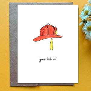Puede incluir: Tarjeta de felicitación con una ilustración de un casco de bombero rojo y una borla amarilla. El texto "You did it!" está escrito en un estilo manuscrito. La tarjeta está sobre un sobre gris, sobre un fondo amarillo.