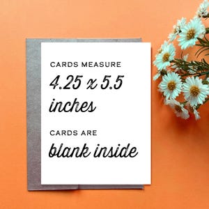 Op de afbeelding: Een witte rechthoekige wenskaart met zwarte tekst die luidt CARDS MEASURE 4.25 x 5.5 inches en CARDS ARE blank inside. De kaart ligt op een grijze envelop tegen een oranje achtergrond, met een bosje witte bloemen ernaast.