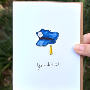 Op de afbeelding: Een witte wenskaart met een aquarelillustratie van een blauwe politiepet met een zilveren badge en een gele kwast. Onder de pet staat de tekst "You did it!" in een eenvoudig, zwart, handgeschreven lettertype.