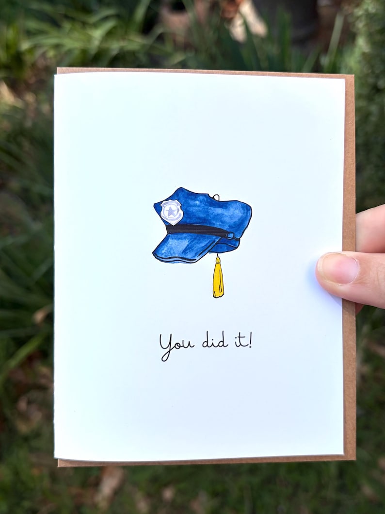 Op de afbeelding: Een witte wenskaart met een aquarelillustratie van een blauwe politiepet met een zilveren badge en een gele kwast. Onder de pet staat de tekst "You did it!" in een eenvoudig, zwart, handgeschreven lettertype.