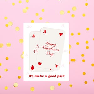 May include: Two playing cards with red suits and the text "Happy Valentine's Day" on a white background. The phrase "We make a good pair" is printed below the cards. The background is pink with gold confetti.