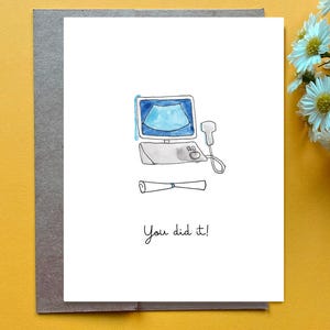 Puede incluir: Una tarjeta de felicitación blanca con una ilustración en acuarela de una máquina de ultrasonido médica con pantalla azul y sonda manual, acompañada de un diploma enrollado y el texto impreso en negro "You did it!" debajo.
