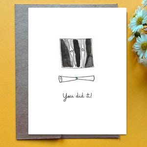 Pode incluir: Um cartão de felicitações de formatura com uma ilustração em aquarela. O cartão apresenta uma imagem de raio-x de ossos, um diploma enrolado e o texto manuscrito "You did it!". O cartão está em um envelope cinza, em um fundo amarelo.