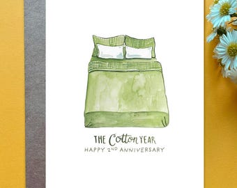 May include: A white greeting card featuring a watercolor illustration of a bed with green bedding and two white pillows. Below the bed, the text reads THE COTTON YEAR HAPPY 2ND ANNIVERSARY in dark gray ink on the card surface.