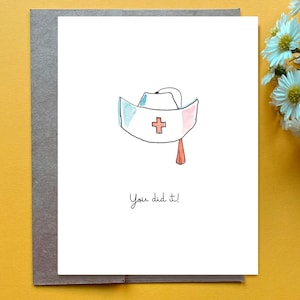Puede incluir: Una tarjeta de felicitación blanca con una ilustración en acuarela de un gorro de enfermería con una cruz roja y una pequeña borla roja. El texto "You did it!" está impreso en tinta negra debajo de la ilustración. La tarjeta está sobre un sobre gris.
