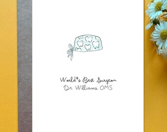 May include: A white greeting card featuring a hand-drawn illustration of a light blue surgical cap with a tooth pattern. Below the illustration, the text reads: World's Best Surgeon, Dr. Williams, OMS. The card is placed on a dark gray envelope.