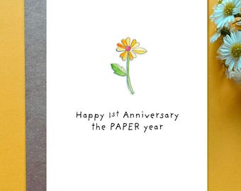 May include: A white greeting card featuring a watercolor illustration of a single yellow and orange daisy with a green stem. Below the flower, the text reads: Happy 1st Anniversary the PAPER year. The card is placed on a dark gray envelope.