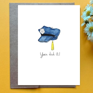 Op de afbeelding: Een witte wenskaart met een aquarelillustratie van een blauwe politiepet met een zilveren badge en een gele kwast aan de zijkant. Onder de pet staat de tekst "You did it!" gedrukt in een eenvoudig, zwart, handgeschreven lettertype.