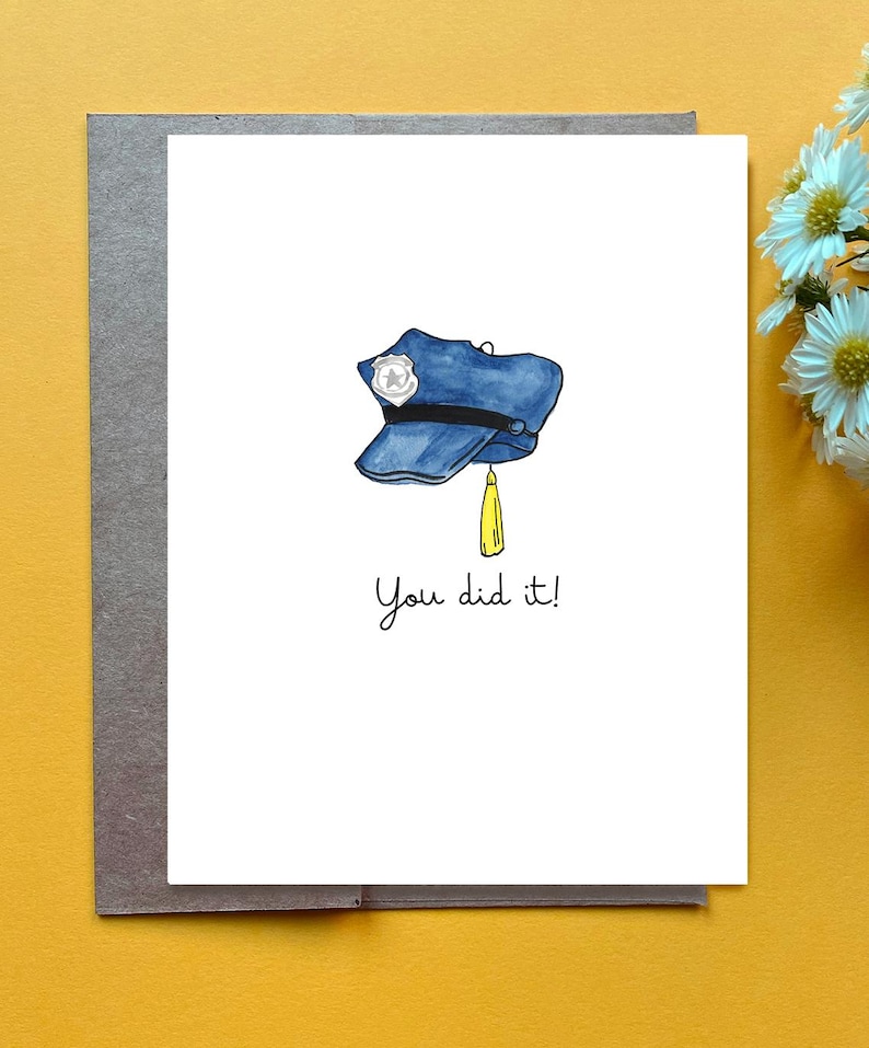 Op de afbeelding: Een witte wenskaart met een aquarelillustratie van een blauwe politiepet met een zilveren badge en een gele kwast aan de zijkant. Onder de pet staat de tekst "You did it!" gedrukt in een eenvoudig, zwart, handgeschreven lettertype.