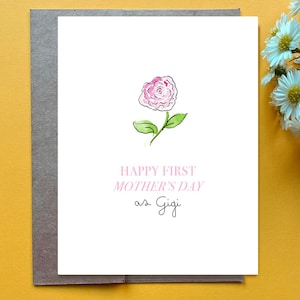 May include: A greeting card with a watercolor illustration of a pink rose and green leaves. The card reads "HAPPY FIRST MOTHER'S DAY as Gigi" in pink and black script. The card is on a gray envelope, set against a yellow background.