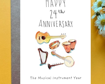 May include: A white greeting card with the text Happy 24th Anniversary and The Musical Instrument Year. The card features watercolor illustrations of a guitar, tambourine, two drums, a harp, a banjo, and a flute in various colors.