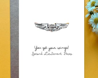 May include: A white greeting card featuring a hand-drawn illustration of silver aviator wings with a central shield emblem. Below the wings, the text reads: You got your wings! Second Lieutenant Perez. The card is placed on a dark gray envelope.