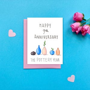 Könnte beinhalten: Eine weiße Grußkarte mit dem Text "HAPPY 9th ANNIVERSARY" und "THE POTTERY YEAR" liegt auf einem rosa Umschlag. Die Karte zeigt Aquarellillustrationen von Vasen und einem schwarzen Stein. Rosa Herzen und Rosen sind verstreut.