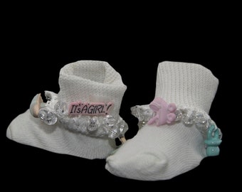 May include: Two white baby socks, each with decorative trim. One sock features a pink label saying "IT'S A GIRL!", the other a pink stroller charm. Both socks have clear crystal-like accents.