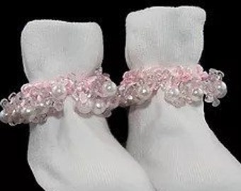 May include: A pair of white baby socks with a pink beaded trim. The socks are made of a soft, ribbed material and feature a delicate, decorative band of pink beads around the ankle.