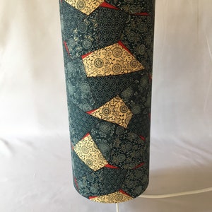 May include: A tall, cylindrical lampshade with a white base. The lampshade is covered in a dark blue fabric with a repeating pattern of gold and red geometric shapes and floral designs.