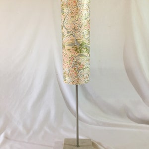 May include: A table lamp with a cylindrical shade featuring a floral pattern in shades of pink, white, and green. The lamp has a silver metal stem and a square concrete base.