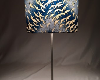 May include: A table lamp with a white and blue patterned lampshade featuring a flock of birds. The lamp has a silver metal stem and a square concrete base.