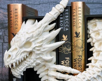 Puede incluir: Un juego de sujetalibros blanco con diseño de dragón que presenta una cabeza de dragón detallada a la izquierda y una cola segmentada a la derecha, colocado entre varios libros de Rebecca Yarros titulados Fourth Wing y Onyx Storm.