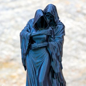 May include: A dark blue, monochromatic figurine depicting two hooded, skeletal figures in a close embrace. The figures are draped in long, flowing robes with visible folds, and the skeletal hands of one figure are wrapped around the other's torso.